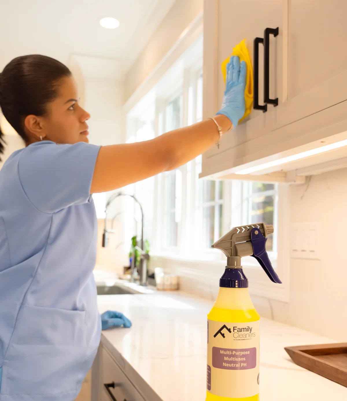 A Family Cleaners team member in gloves wiping white kitchen cabinet doors, with a branded spray bottle on the counter
