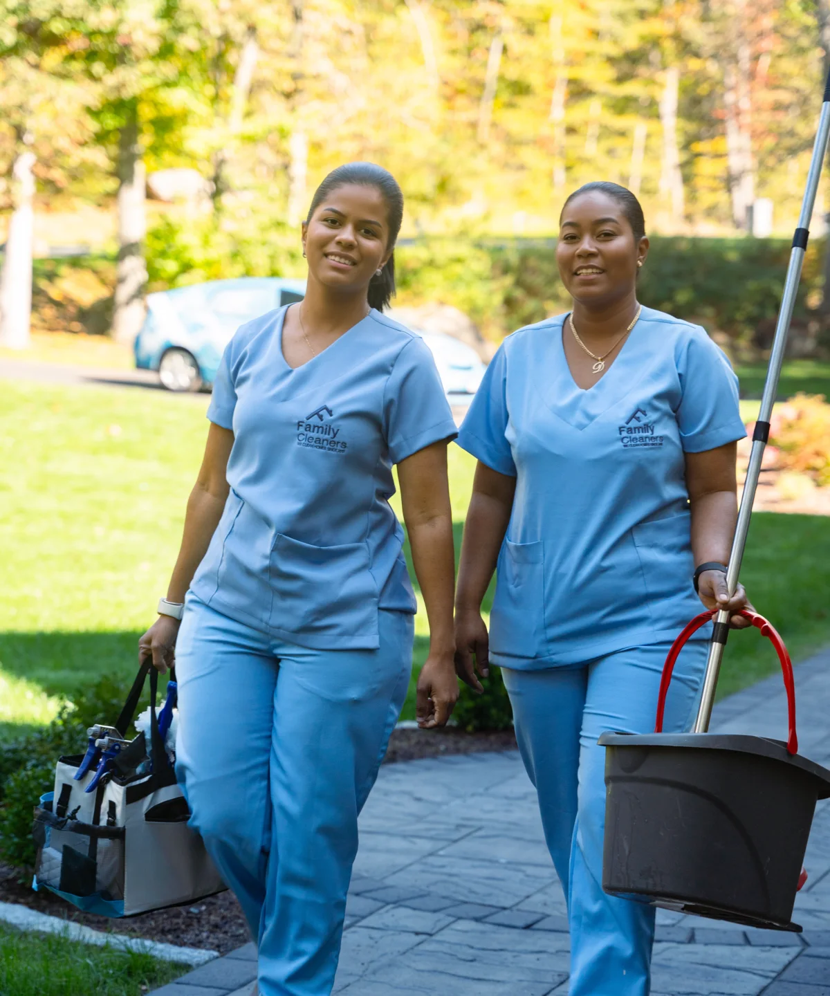 Two Family Cleaners team members in blue uniforms walking up a garden path carrying a cleaning caddy, a bucket and a mop